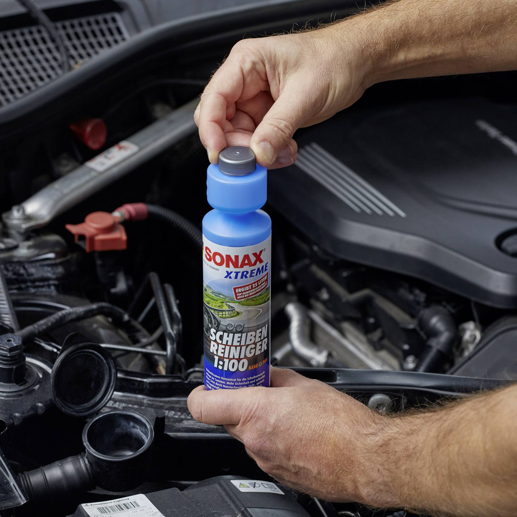 Une personne tient une bouteille de nettoyant pour vitres portant l'inscription 'Sonax Xtreme' au-dessus d'un compartiment moteur ouvert.