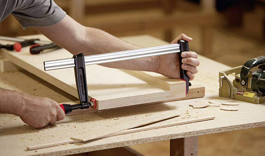 Une personne fixe des morceaux de bois avec un serre-joint dans un atelier. Des copeaux de bois sont éparpillés sur la table et une scie est visible.