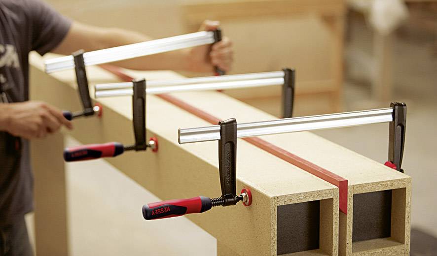 Une personne utilise plusieurs serre-joints pour maintenir des pièces de bois fixées sur un établi. La scène illustre un travail de précision dans un atelier.