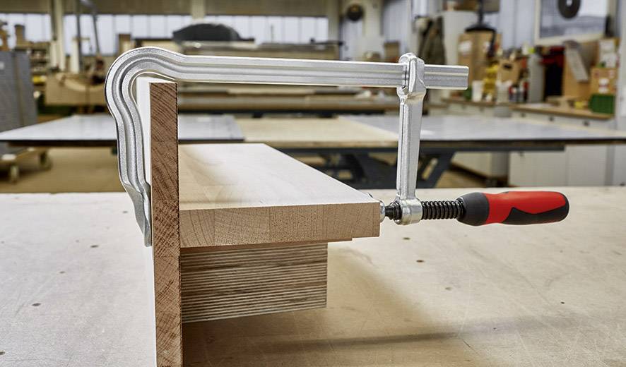 Un morceau de bois est fixé à un bord de table à l'aide de deux serre-joints. En arrière-plan, on aperçoit un atelier avec des surfaces de travail et des outils.