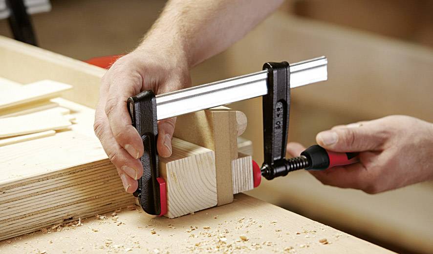 Deux mains utilisent un serre-joint pour maintenir deux morceaux de bois fixés sur une table. Des copeaux de sciure sont dispersés autour des morceaux de bois.