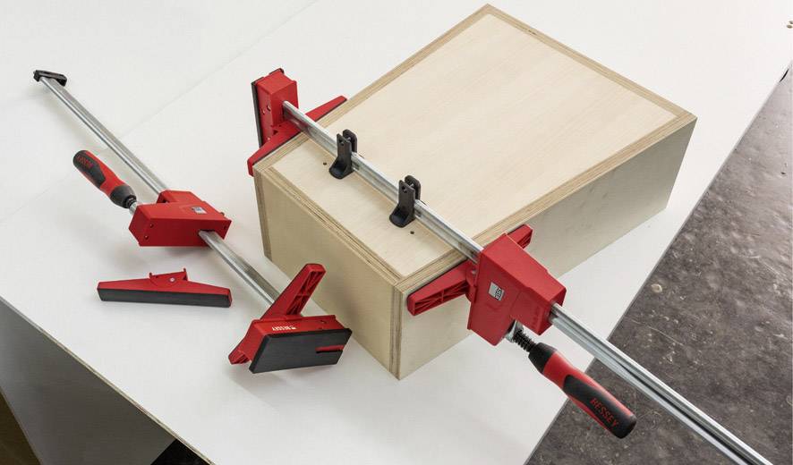 Une caisse en bois est fixée à l'aide de plusieurs serre-joints. Les serre-joints ont des poignées rouges et des tiges métalliques qui assurent la stabilité.