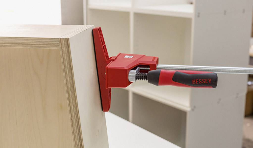 Un système de serrage avec une poignée rouge maintient deux pièces de bois ensemble, probablement pour un assemblage ou des travaux de menuiserie dans un atelier.