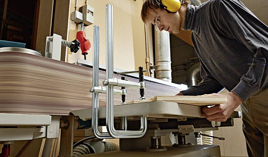 Un homme utilise une meuleuse dans un atelier. Il porte des protections auditives et travaille une planche en bois.