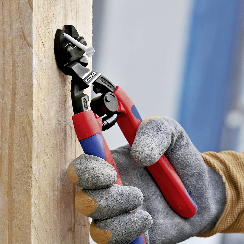 Une personne portant des gants de travail coupe un clou d'une planche de bois à l'aide d'une pince rouge.