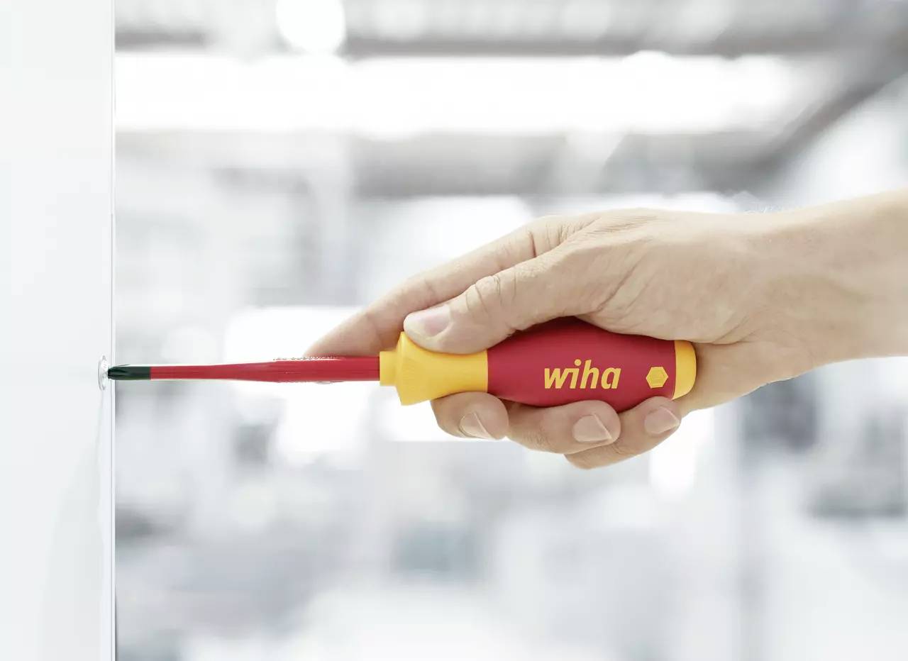 Une main tient un tournevis rouge avec une poignée jaune pendant qu'il visse une vis dans une surface blanche.