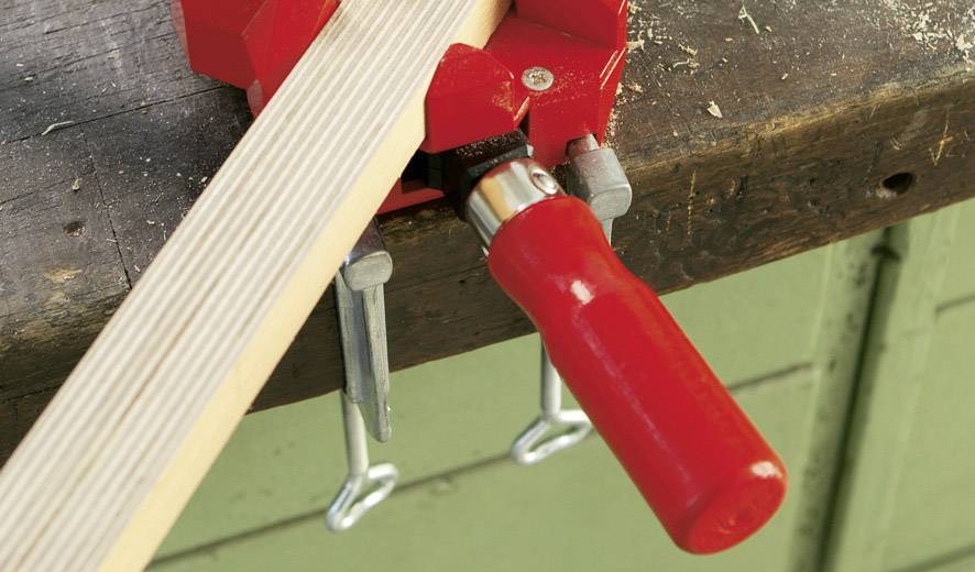 Une poutre en bois est maintenue par un serre-joint rouge sur un établi. Celui-ci sert à immobiliser la poutre pendant le travail.