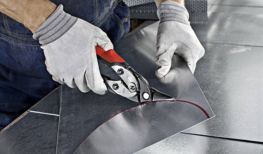 Une personne porte des gants de travail et coupe un morceau de tôle métallique avec des cisailles dans un atelier.