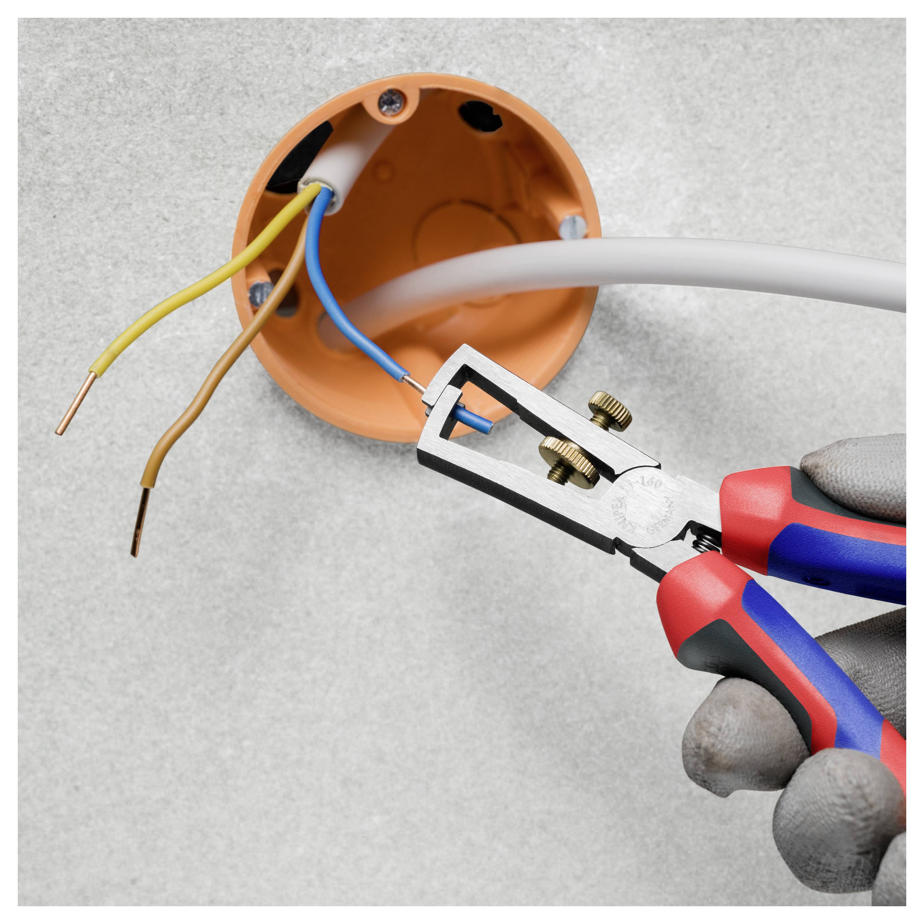 Des mains gantées utilisent une pince à dénuder pour enlever l'isolant d'un fil bleu provenant d'une boîte de jonction orange ronde avec des fils électriques exposés.