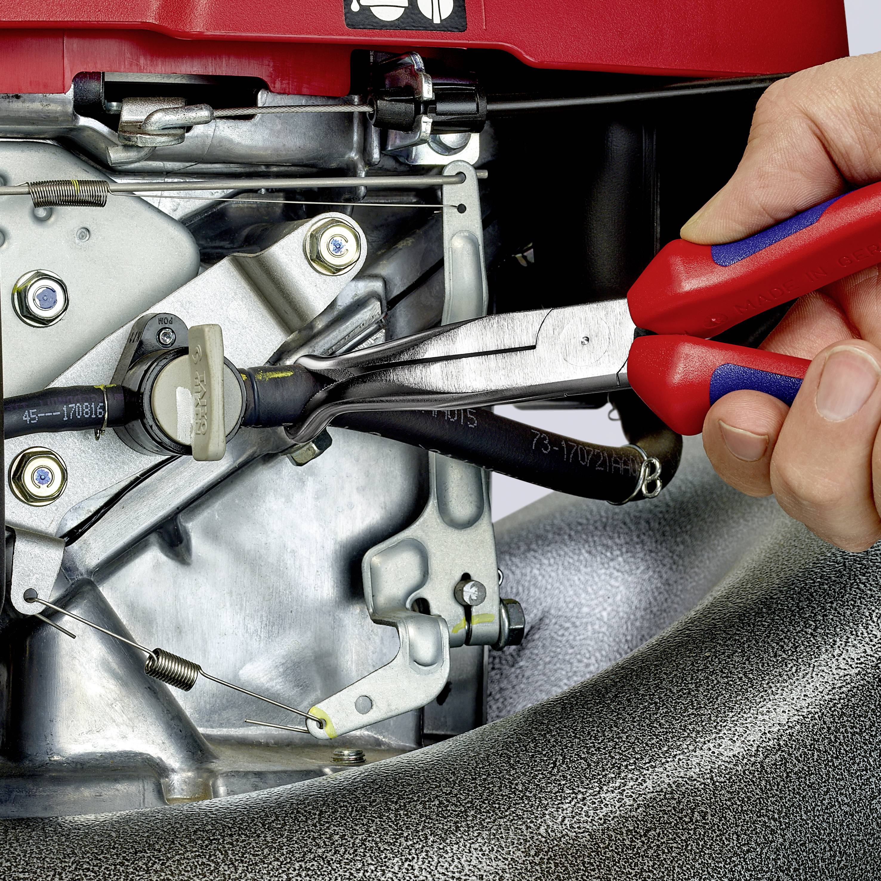 Une main tient une pince et ajuste les câbles d'un moteur de tondeuse à gazon. Des pièces mécaniques du moteur sont visibles.