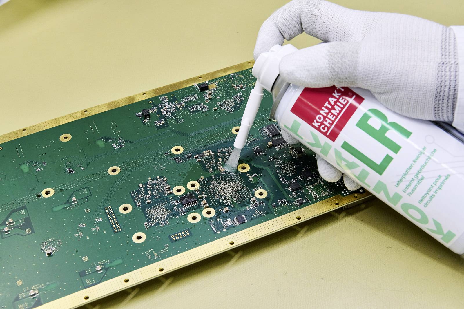 Une main gantée de blanc vaporise un circuit imprimé avec un spray de nettoyage. Le circuit est recouvert de composants verts et dorés.