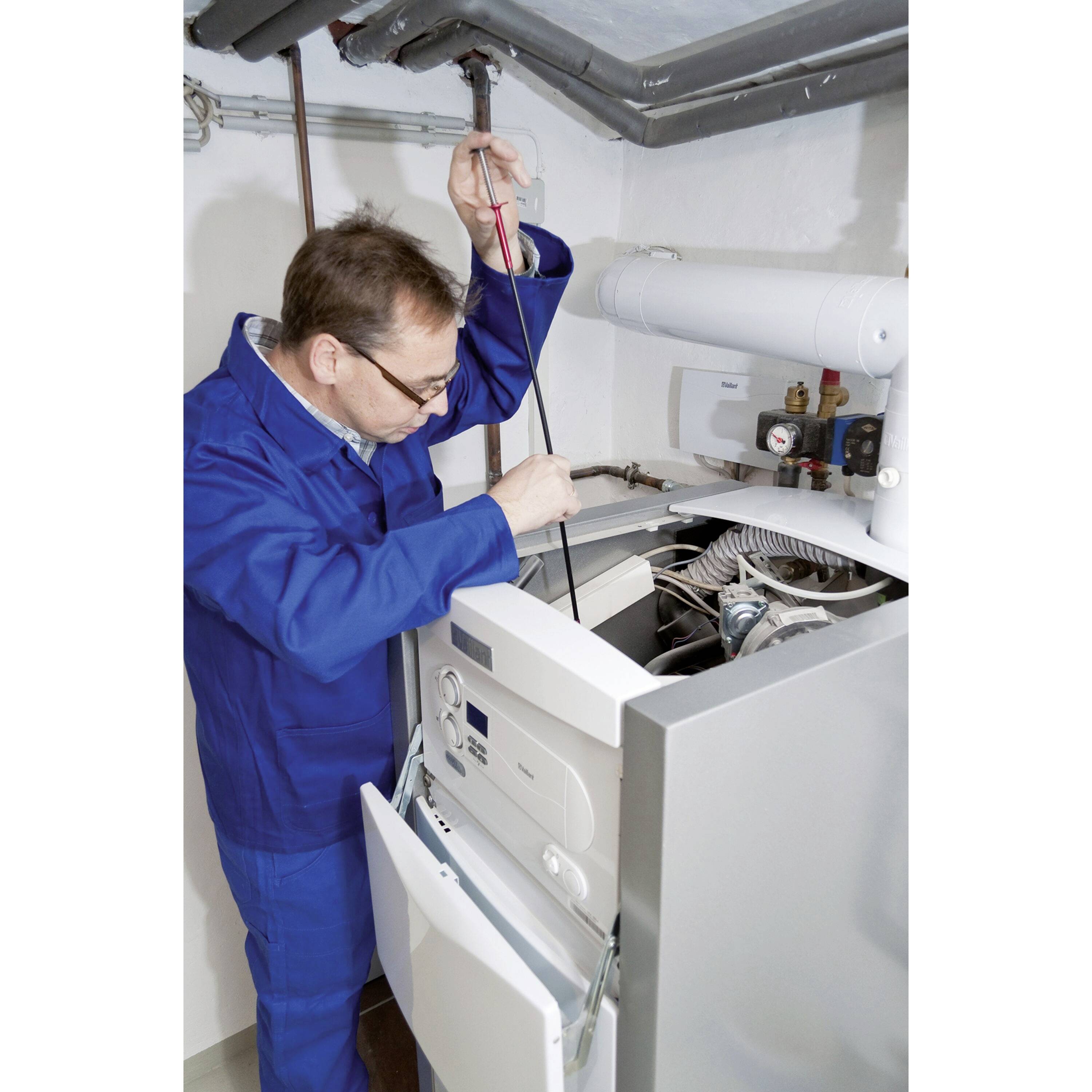 Un technicien en combinaison bleue vérifie l'intérieur d'un système de chauffage avec un outil dans une salle technique.