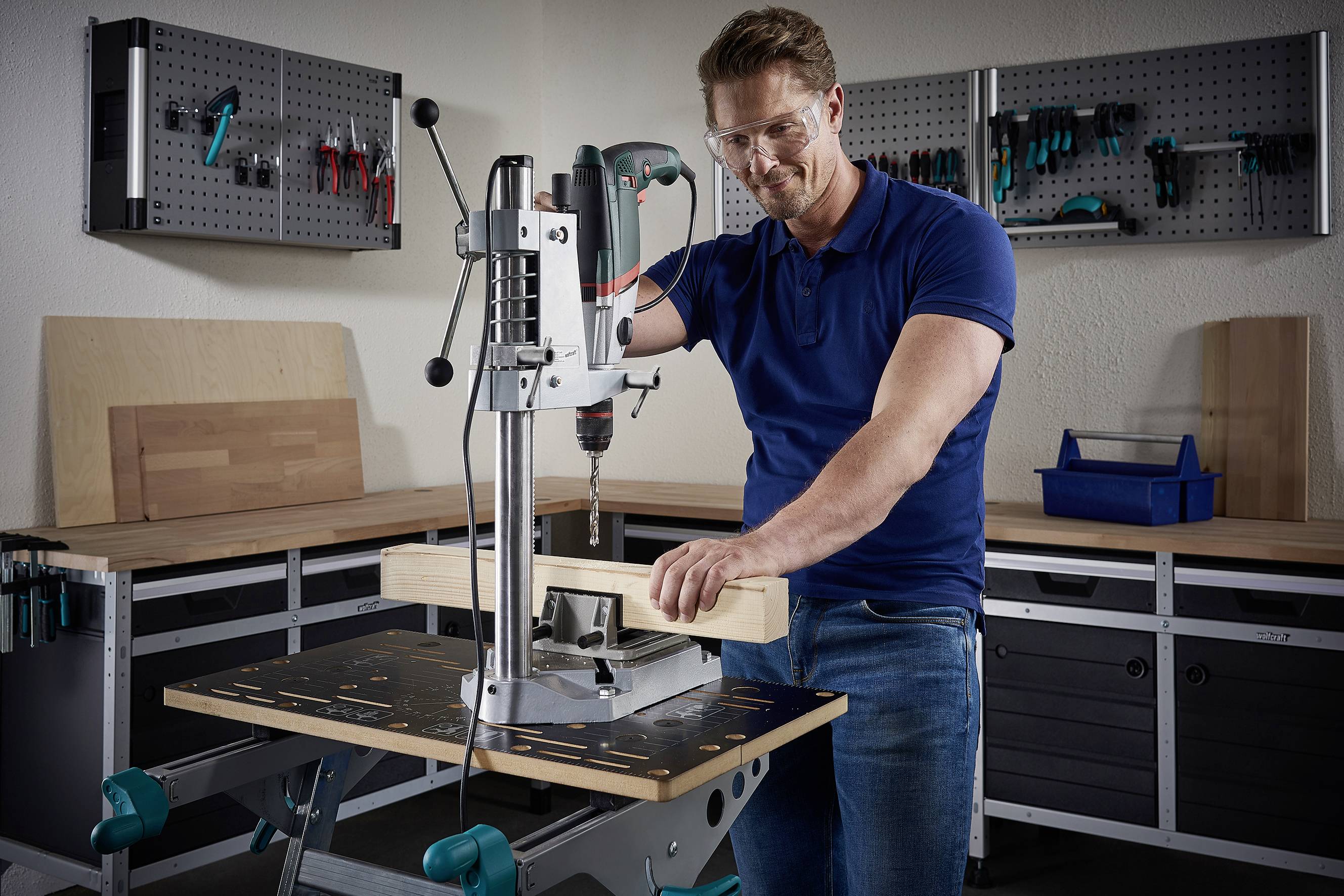 Un homme vêtu de bleu utilise une perceuse à colonne pour travailler une pièce de bois dans un atelier. Des outils sont visibles en arrière-plan.
