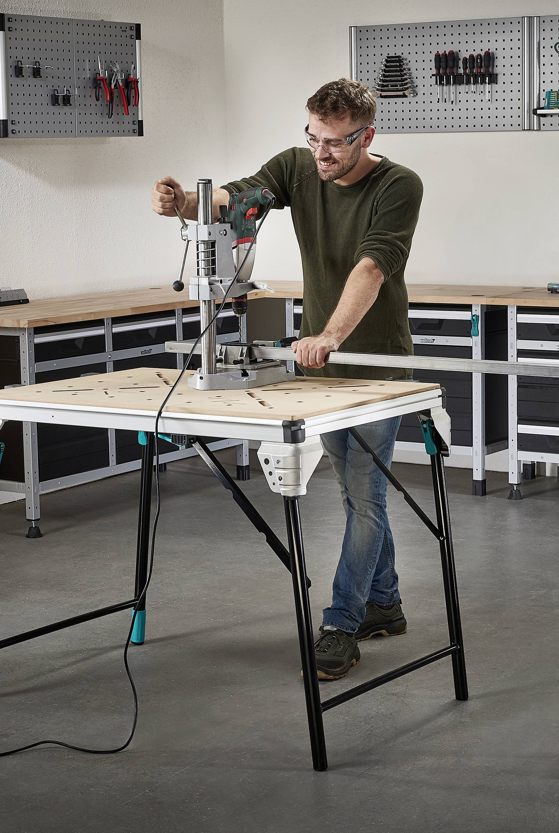 Une personne travaillant avec une perceuse sur une table d'assemblage dans un atelier. Des outils sont visibles sur le mur en arrière-plan.