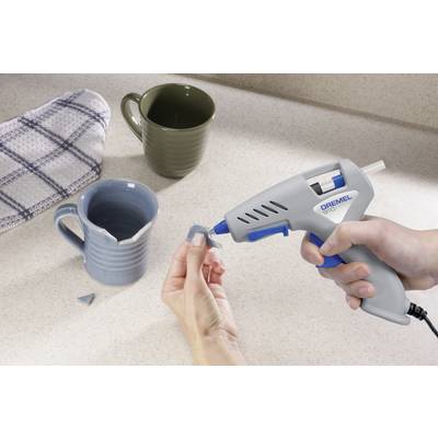 Une personne répare une tasse bleue cassée à l'aide d'un pistolet à colle chaude sur un plan de travail de cuisine. En arrière-plan, deux tasses sont visibles.
