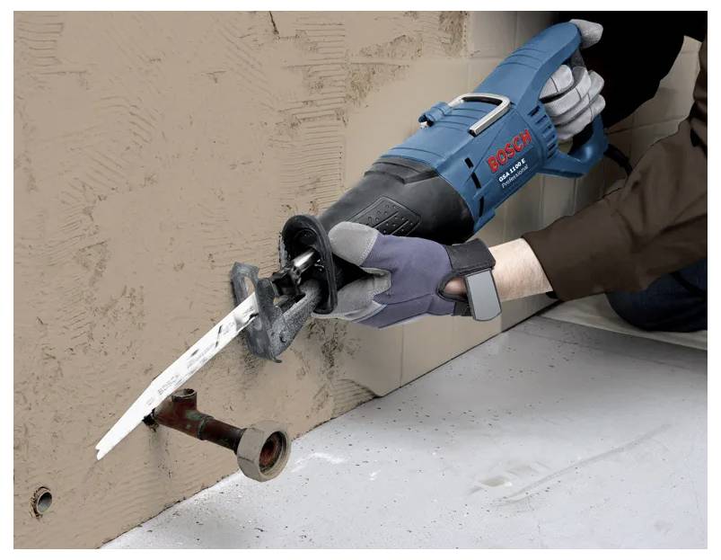 Une personne utilise une scie alternative Bosch pour couper à travers un mur brun, ciblant un tuyau métallique. La personne porte des gants pour des raisons de sécurité.
