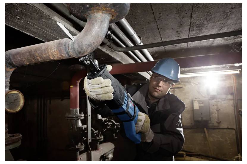 Un opérateur portant un équipement de protection utilise un outil électrique pour couper un tuyau métallique dans un environnement industriel faiblement éclairé.