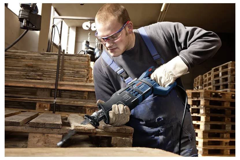 Un homme portant des lunettes de sécurité et des gants utilise une scie alternative pour couper des palettes en bois dans un environnement d'atelier.