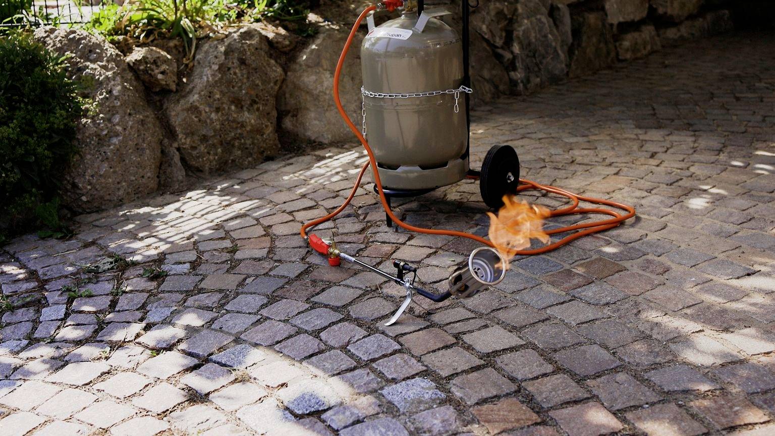 Bouteille de gaz avec un tuyau en feu sur un sol pavé, entourée d'un mur en pierre naturelle. Danger visible dû aux flammes.