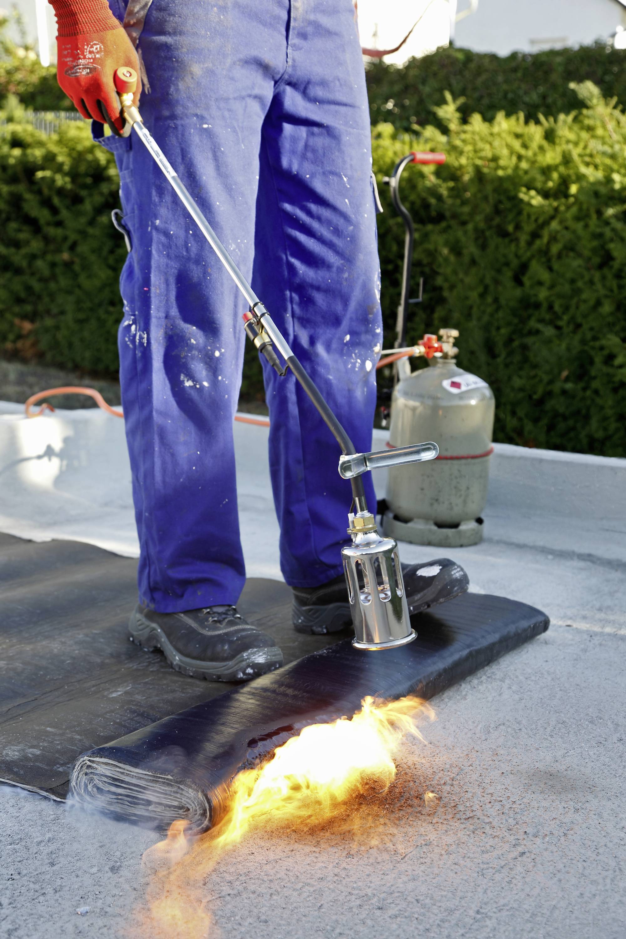 Ouvrier portant un pantalon bleu et des gants rouges utilisant un chalumeau pour souder une membrane de bitume sur du béton, bouteille de gaz en arrière-plan.