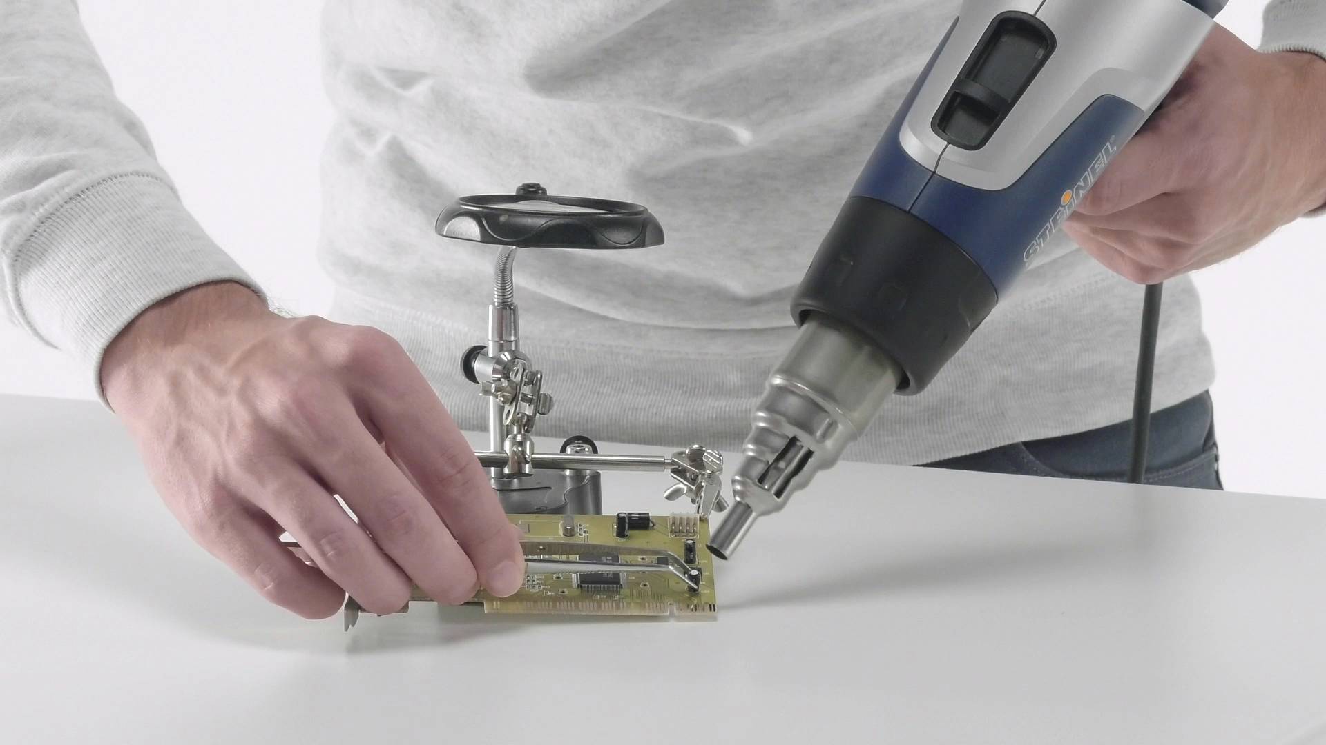 Une personne soude une carte de circuit imprimé avec un pistolet à air chaud. Elle maintient la carte sur un support, tandis que l'appareil est tenu dans l'autre main.