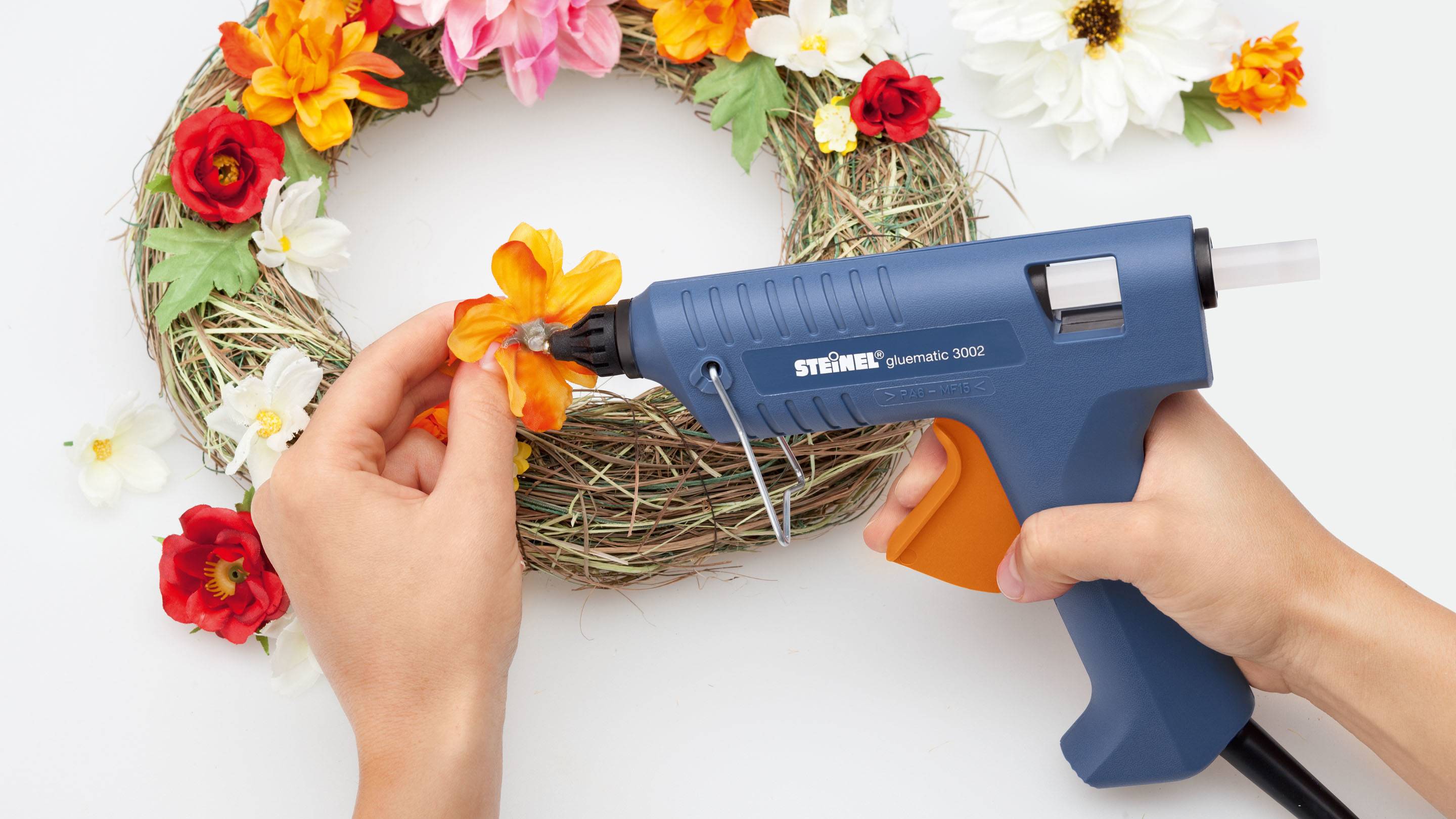 Une personne fixe une fleur orange à l'aide d'un pistolet à colle sur une couronne faite de branches et de fleurs. Une décoration festive est en cours de création.