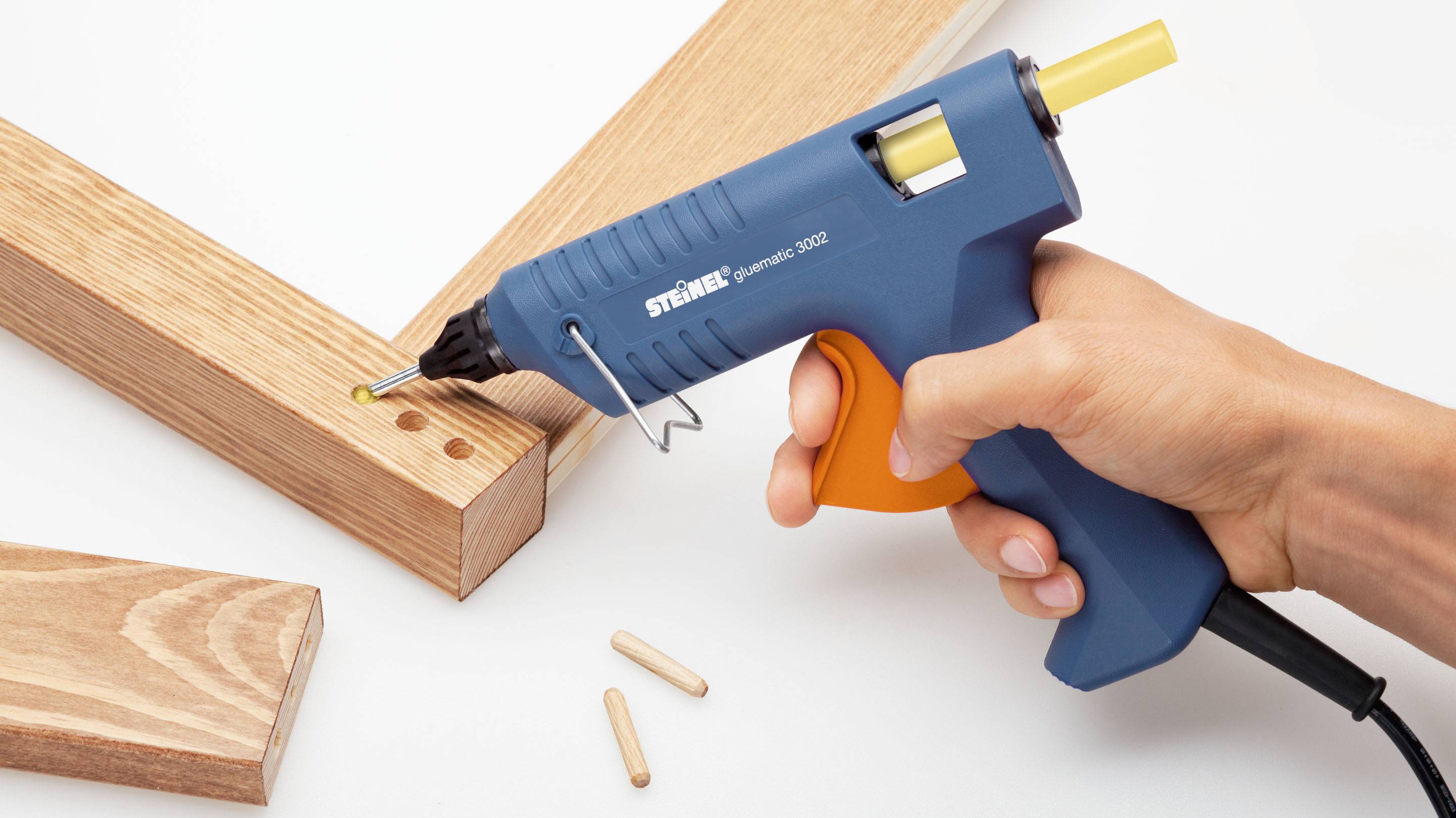 Une main tient un pistolet à colle chaude bleu avec un bâton de colle jaune et colle deux morceaux de bois l'un à l'autre.