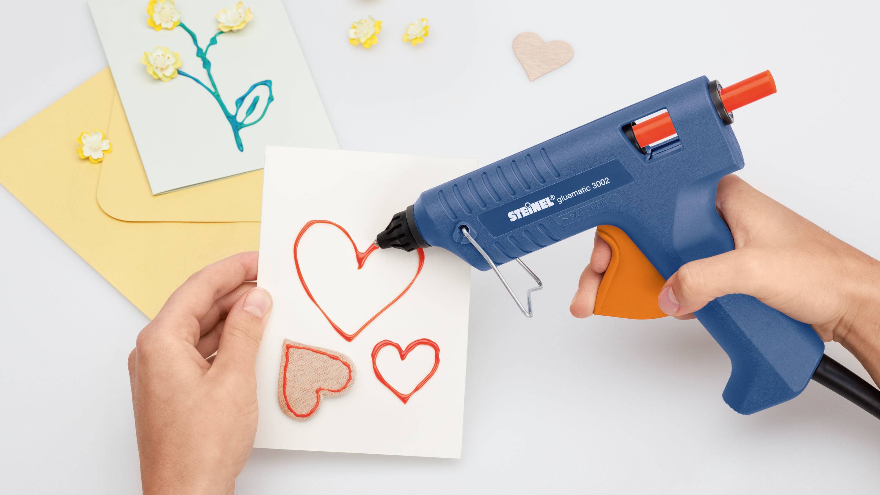 Une personne utilise un pistolet à colle chaude pour coller des cœurs tricolores sur une carte, entourée de matériaux de bricolage.