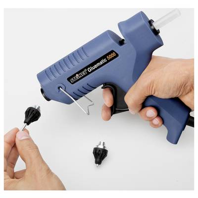 Des mains utilisant un pistolet à colle chaude bleu étiqueté « Steinel Gluematic 5000 » pour appliquer de la colle sur une petite buse noire sur une surface blanche.