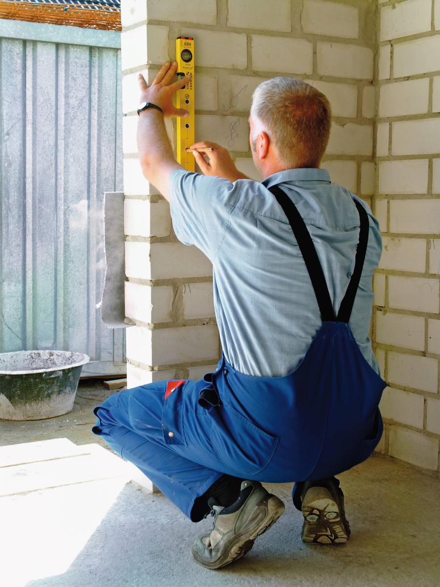 Un artisan en tenue de travail bleue vérifie la verticalité d'un mur en maçonnerie avec un niveau à bulle dans un bâtiment en construction.
