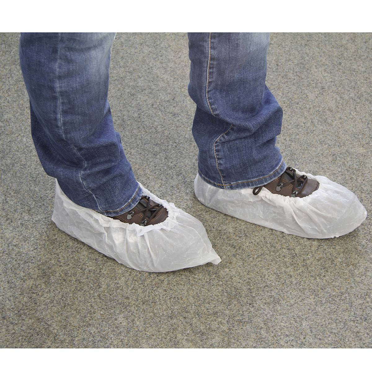 Une personne porte un jean bleu et des chaussures brunes avec des sur-chaussures blanches sur un tapis gris.