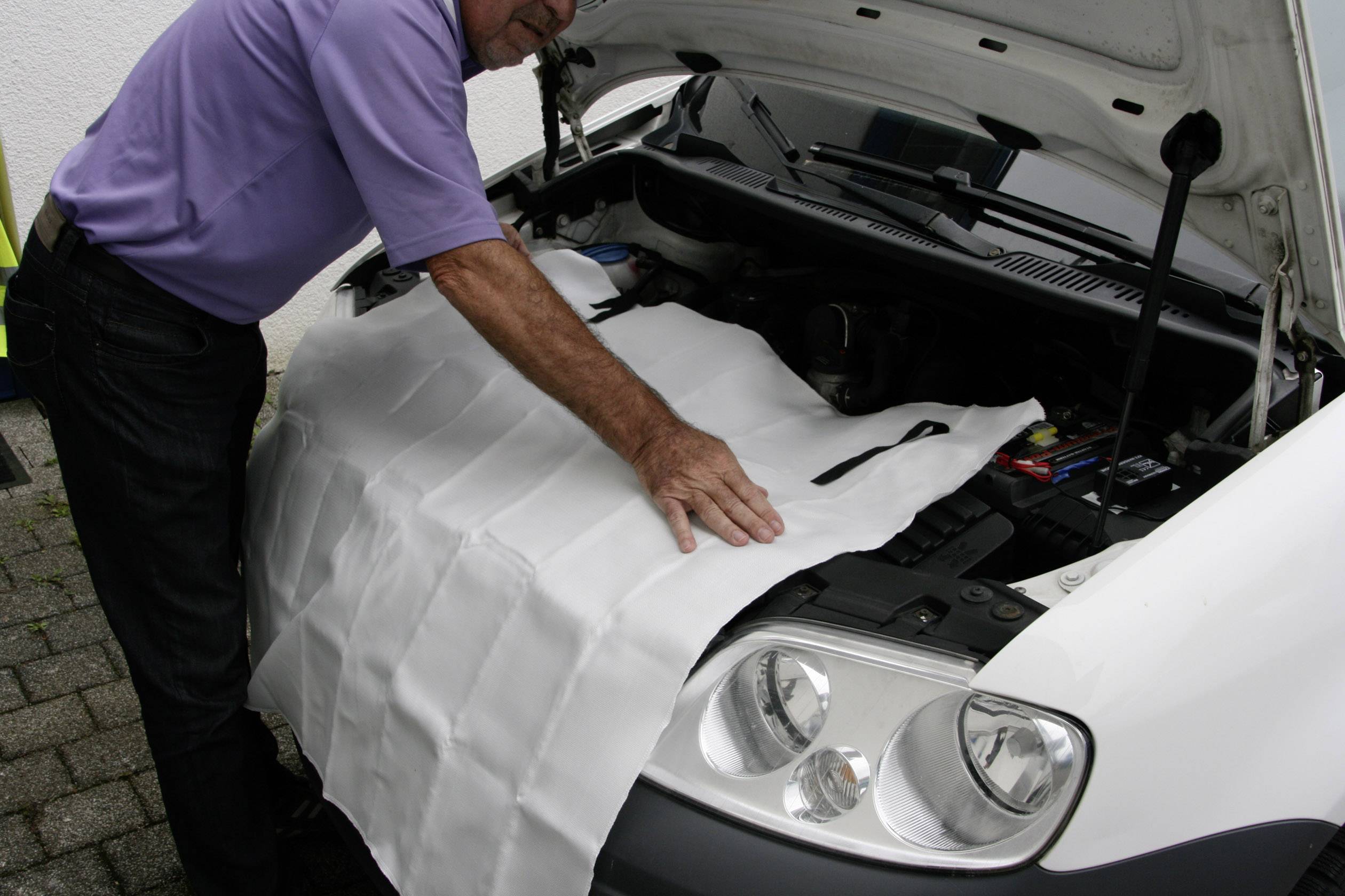 Une personne étale une couverture de protection sur le compartiment moteur d'une voiture. Le capot est ouvert.