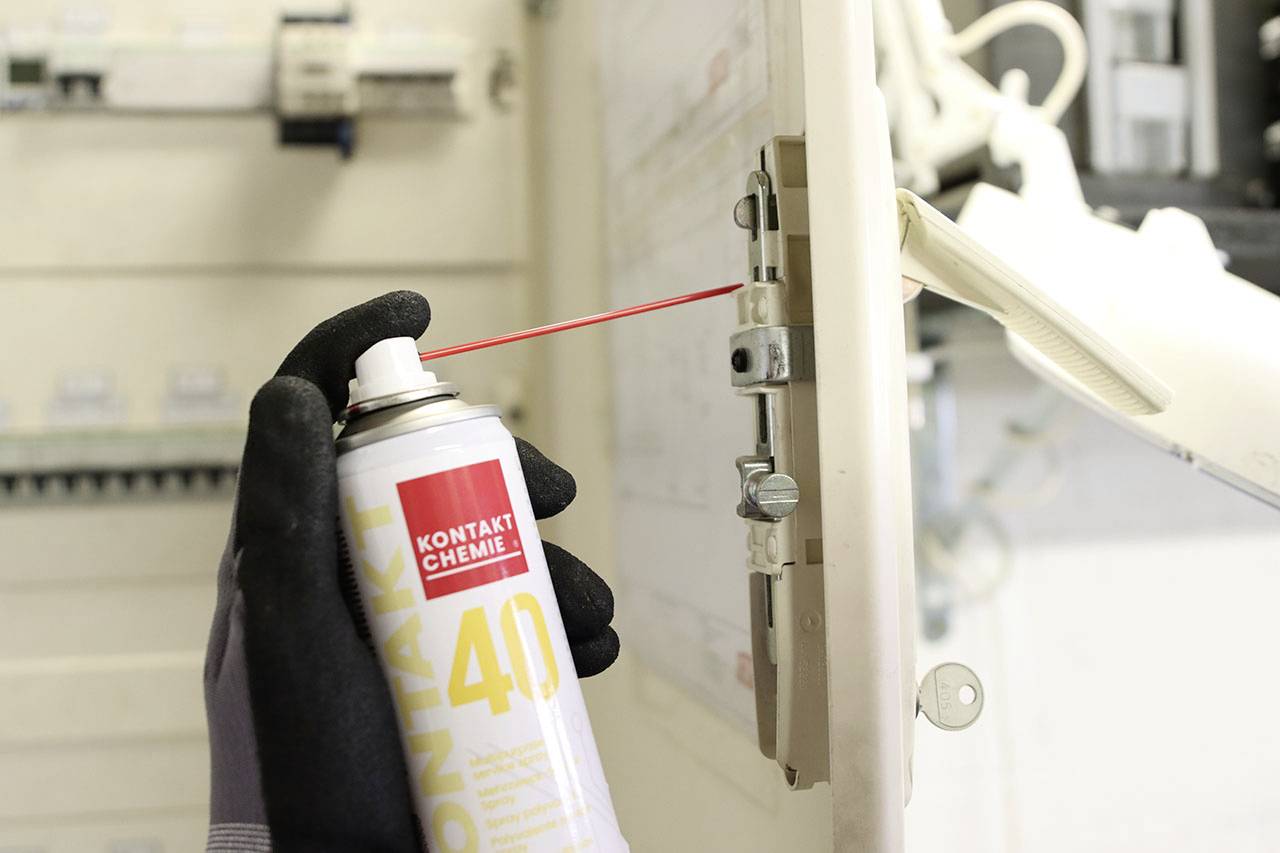 Une personne utilise un spray pour entretenir des parties d'un armoire électrique. L'étiquette de l'aérosol indique 'Kontakt Chemie'.