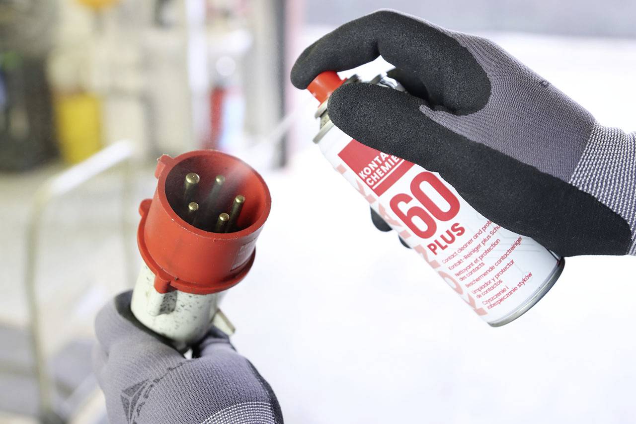 'Gros plan d'une personne portant des gants, tenant un boîtier électrique rouge et vaporisant de l'huile de lubrification d'un bidon.'