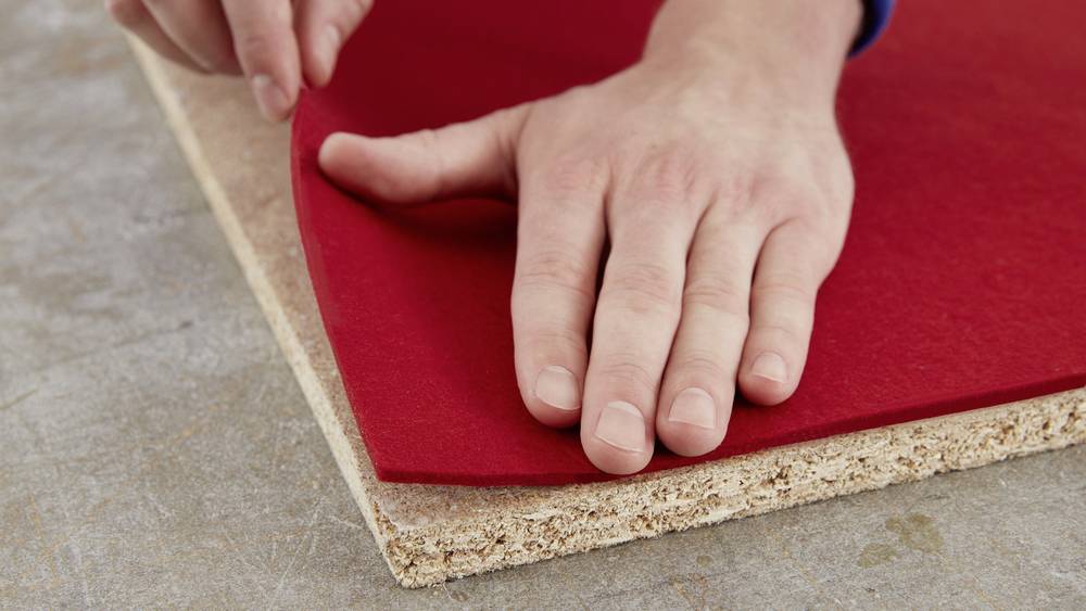 Une main dépose un morceau de feutre rouge sur une planche de particules, probablement pour un travail artisanal ou de construction.