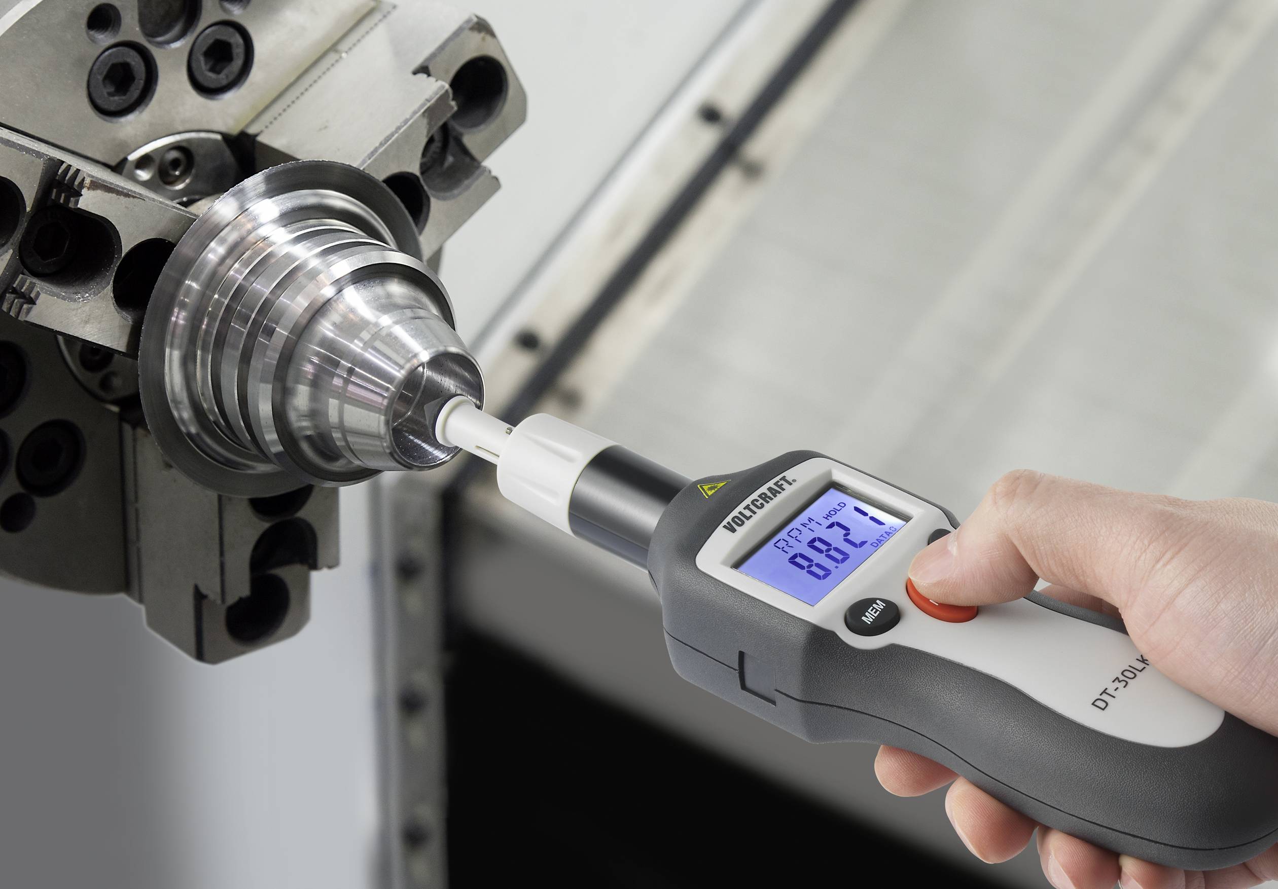 Une main tient un appareil de contrôle et mesure le battement d'une pièce métallique usinée, fixée dans une machine.