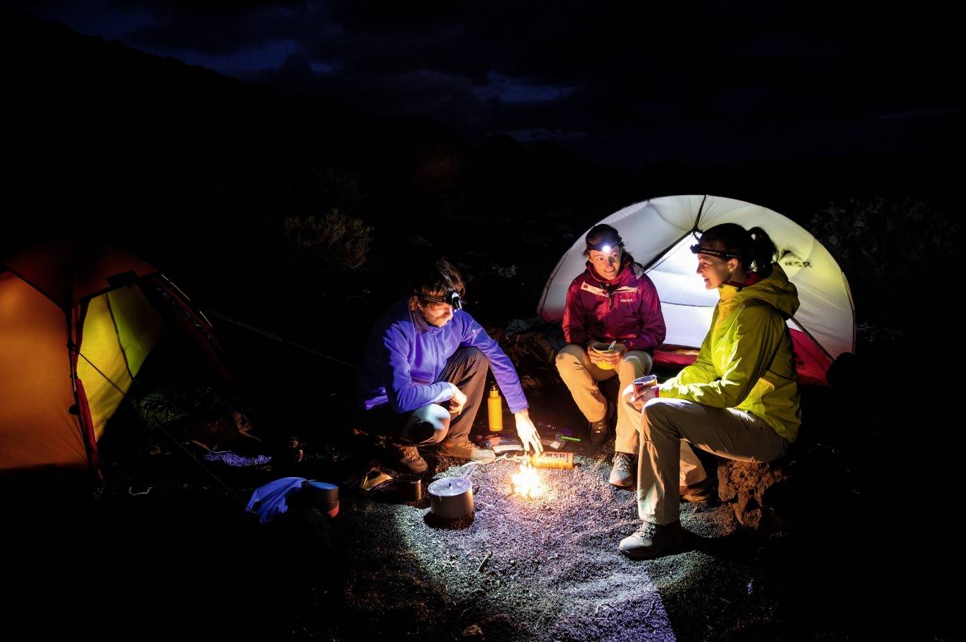 Trois personnes sont assises autour d'un feu de camp dans une forêt sombre, devant deux tentes. Elles portent des vêtements de plein air et profitent de la nuit.