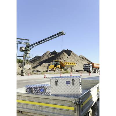 Un grand tas de sable sur un chantier de construction, avec un camion et un engin de chantier en arrière-plan. Au premier plan, une remorque ouverte avec des caisses est visible.