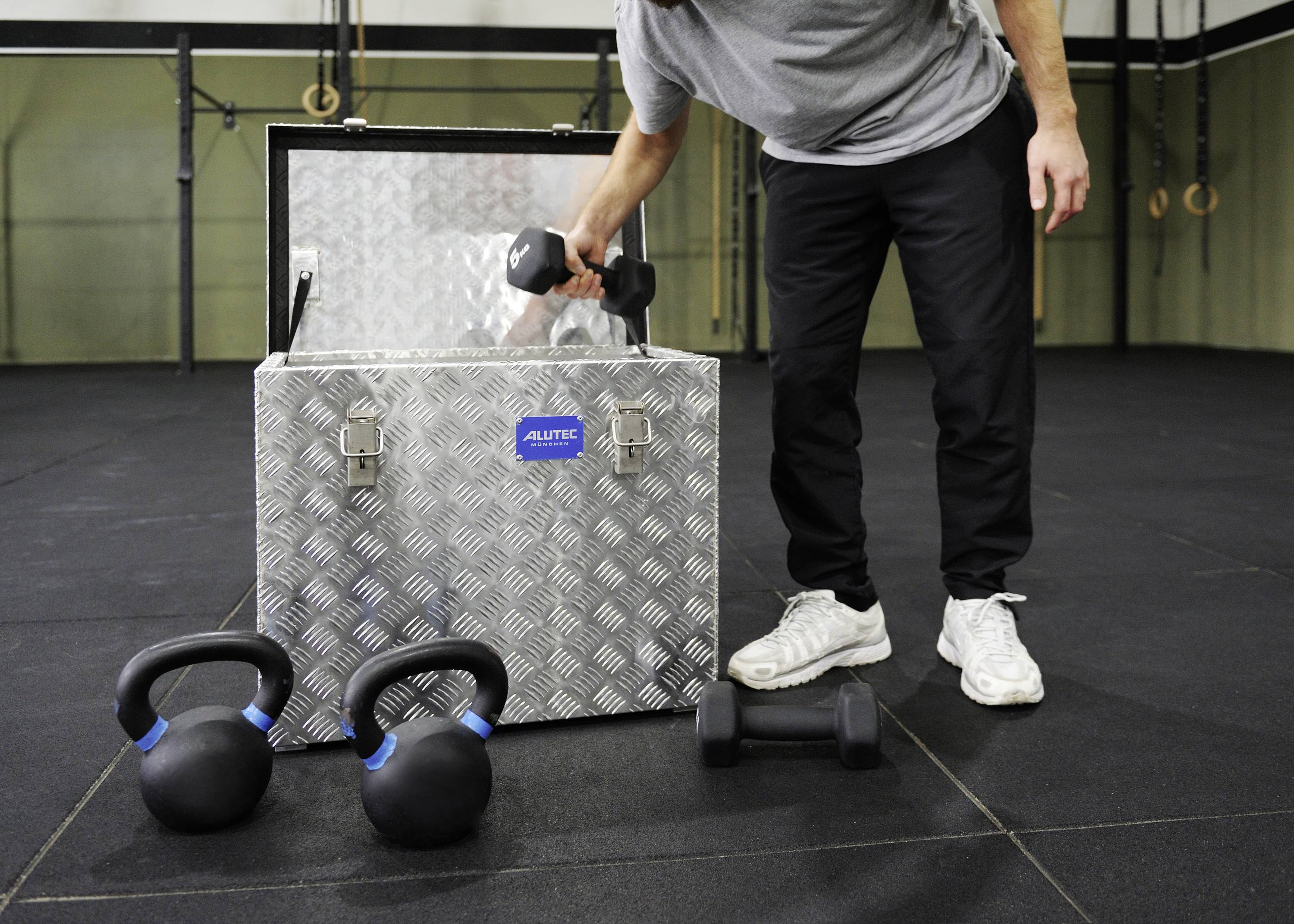 Une personne soulève un haltère d'une boîte métallique dans une salle de sport. Des kettlebells et un autre haltère sont posés sur le sol.