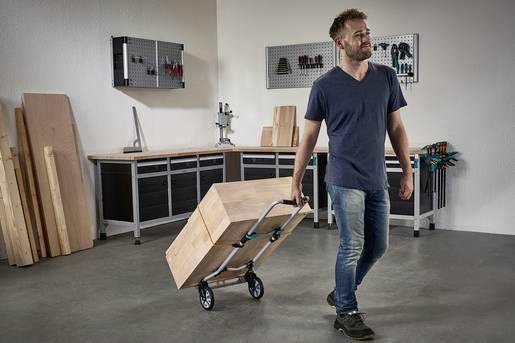 Un homme tire un chariot en bois dans un atelier. À l'arrière-plan, on peut voir des outils accrochés au mur et des planches de bois.