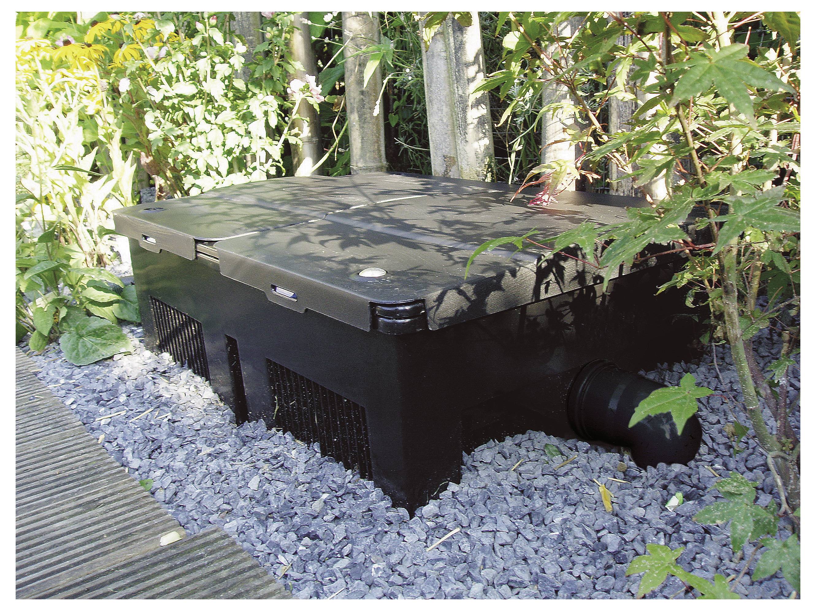 Un boîtier de filtre d'étang noir entouré de verdure et placé sur du gravier à côté d'une terrasse en bois, avec des tuyaux visibles sur son côté.