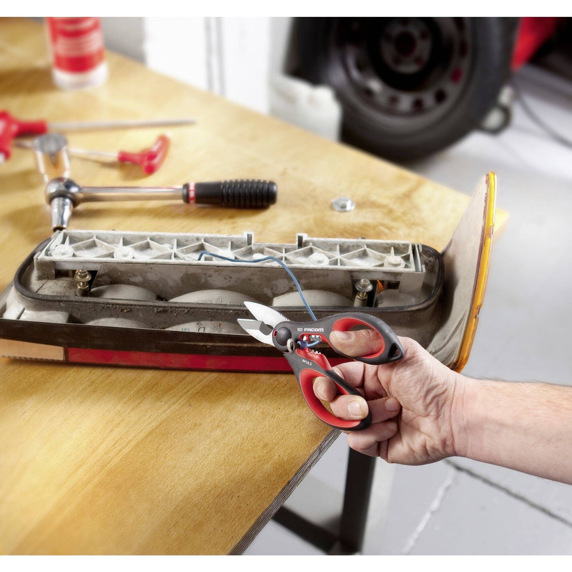 Une main tient une pince rouge et coupe un câble bleu relié à une lampe de voiture sur une table en bois. Des outils sont visibles en arrière-plan.