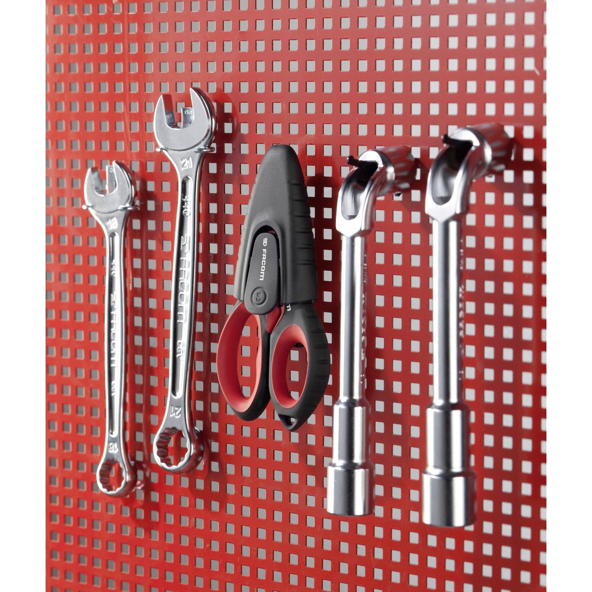 Des clés à molette, des ciseaux et des douilles sont suspendus soigneusement à un mur rouge perforé, dans un atelier ou un garage.