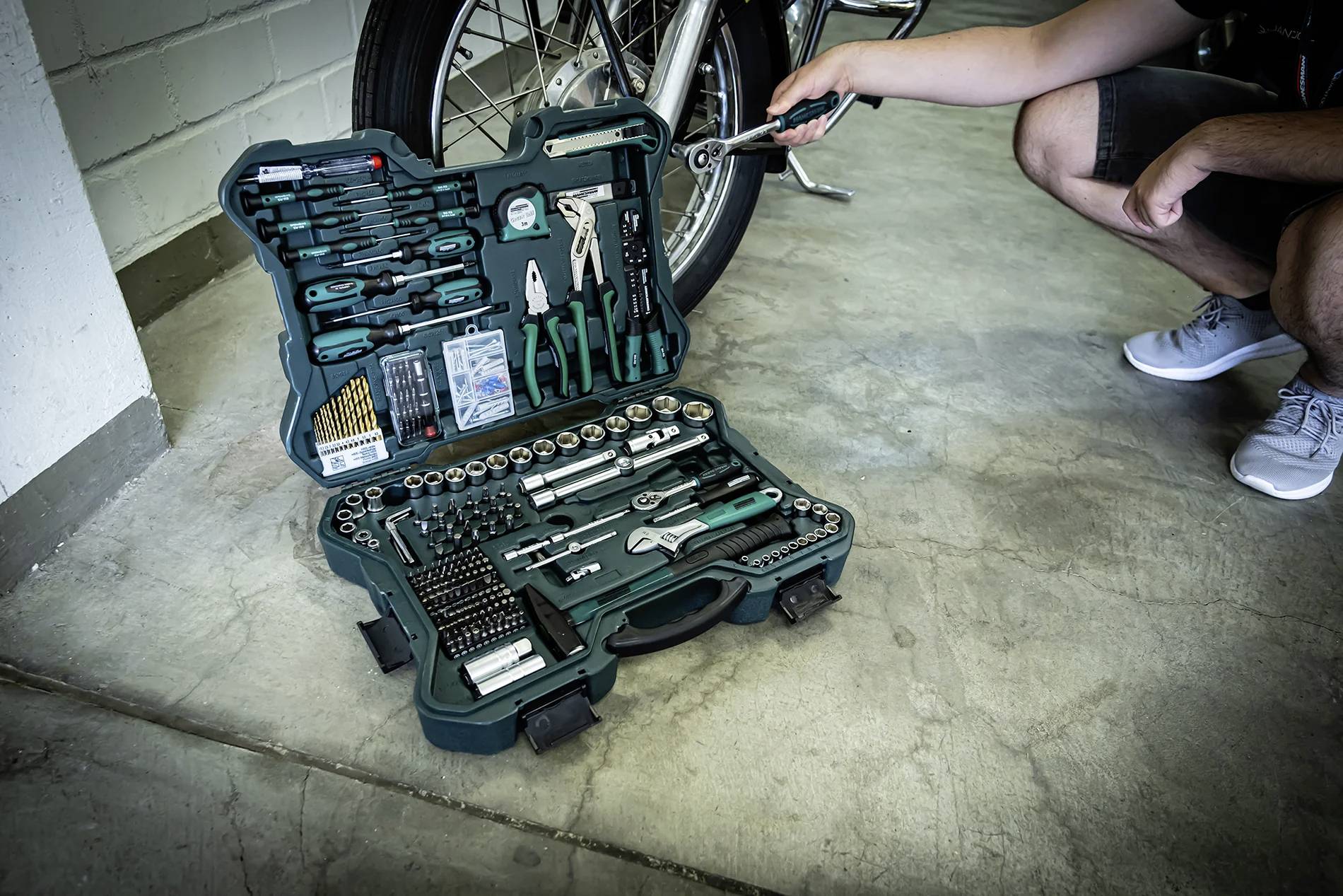 Une boîte à outils verte contenant divers outils manuels, dont des pinces, une clé et un tournevis, est ouverte sur un sol en béton à l'intérieur d'un garage.