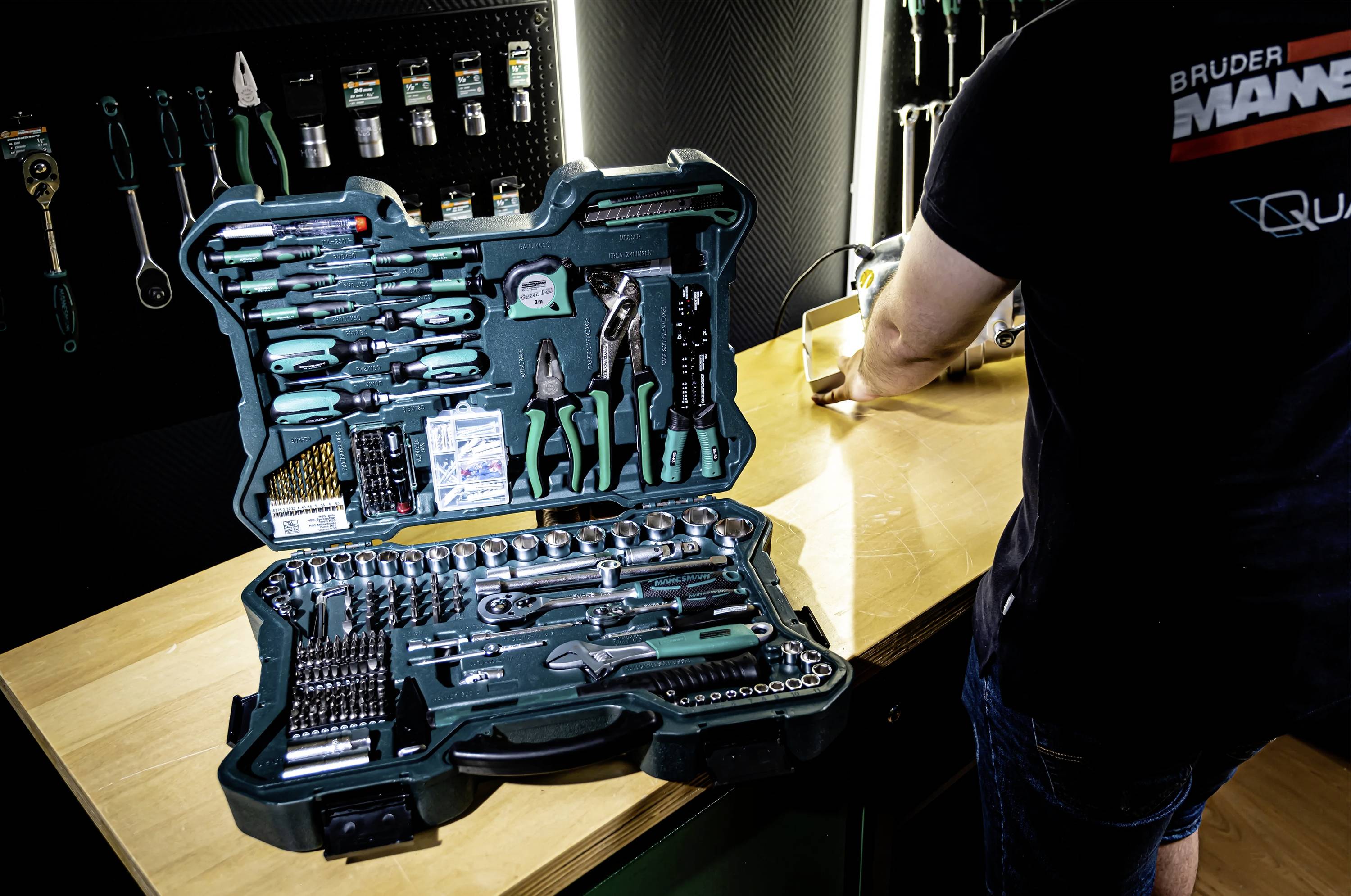 Une boîte à outils ouverte contenant divers outils manuels et accessoires sur une table en bois. Une personne travaille à droite, de dos.