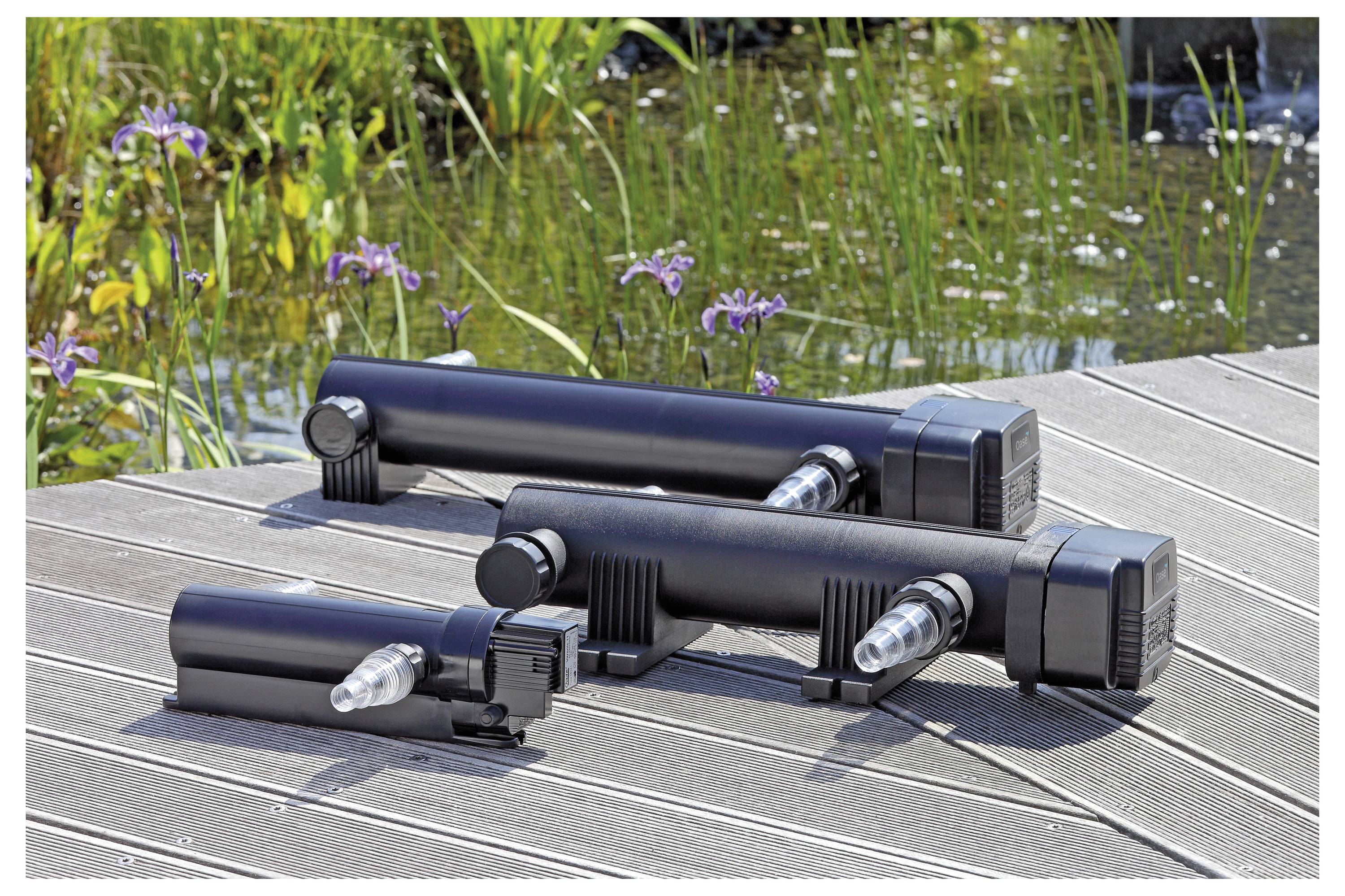 Trois clarificateurs UV noirs de différentes tailles sont placés sur une terrasse en bois à côté d'un étang avec des fleurs violettes en fleur.