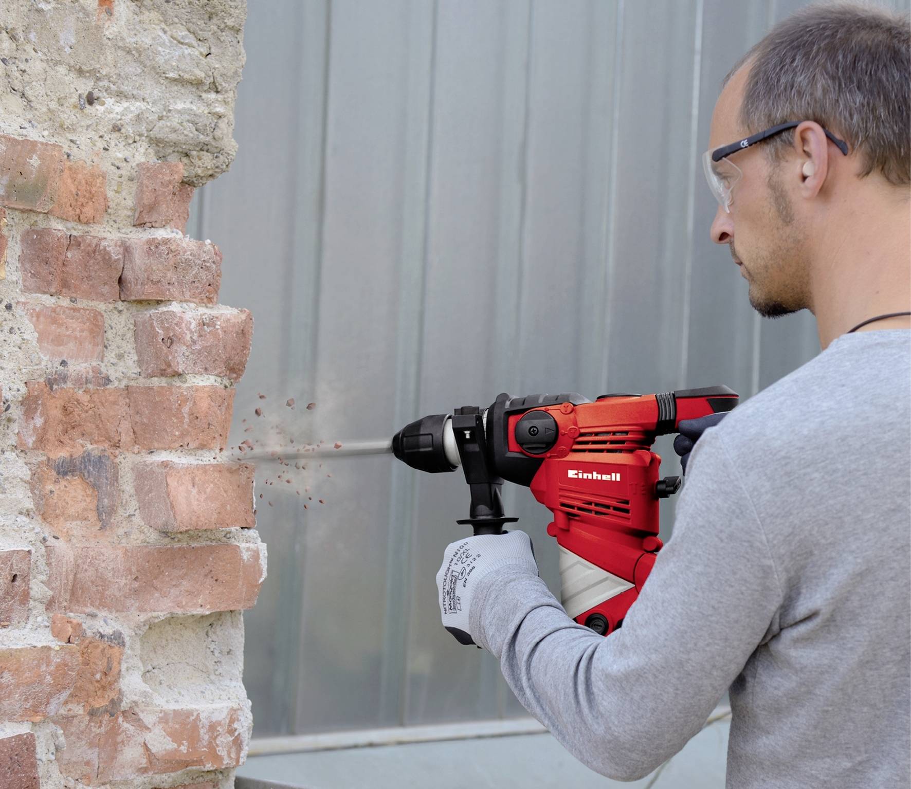 Une personne fore un mur en brique avec un marteau-perforateur rouge. Elle porte des lunettes de protection et des gants.