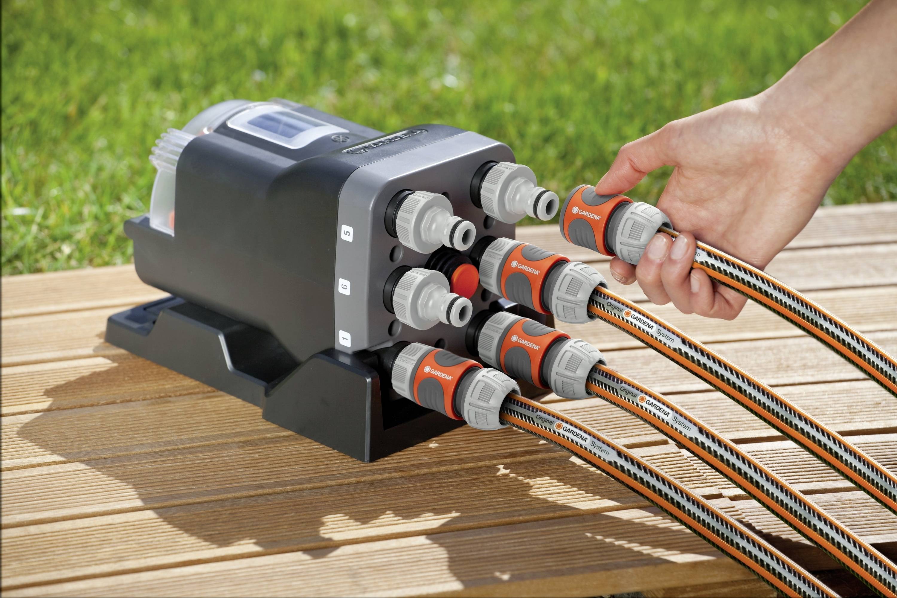 Une main raccorde plusieurs tuyaux d'arrosage à une vanne d'irrigation sur une terrasse en bois, avec de l'herbe visible en arrière-plan.