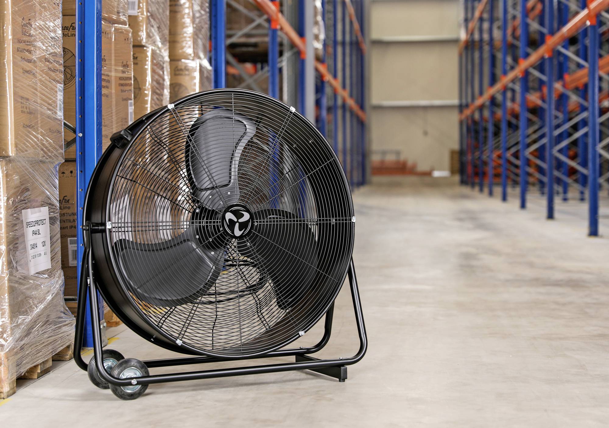 Un grand ventilateur industriel est posé au sol d'un entrepôt, à côté de caisses empilées et d'étagères hautes.