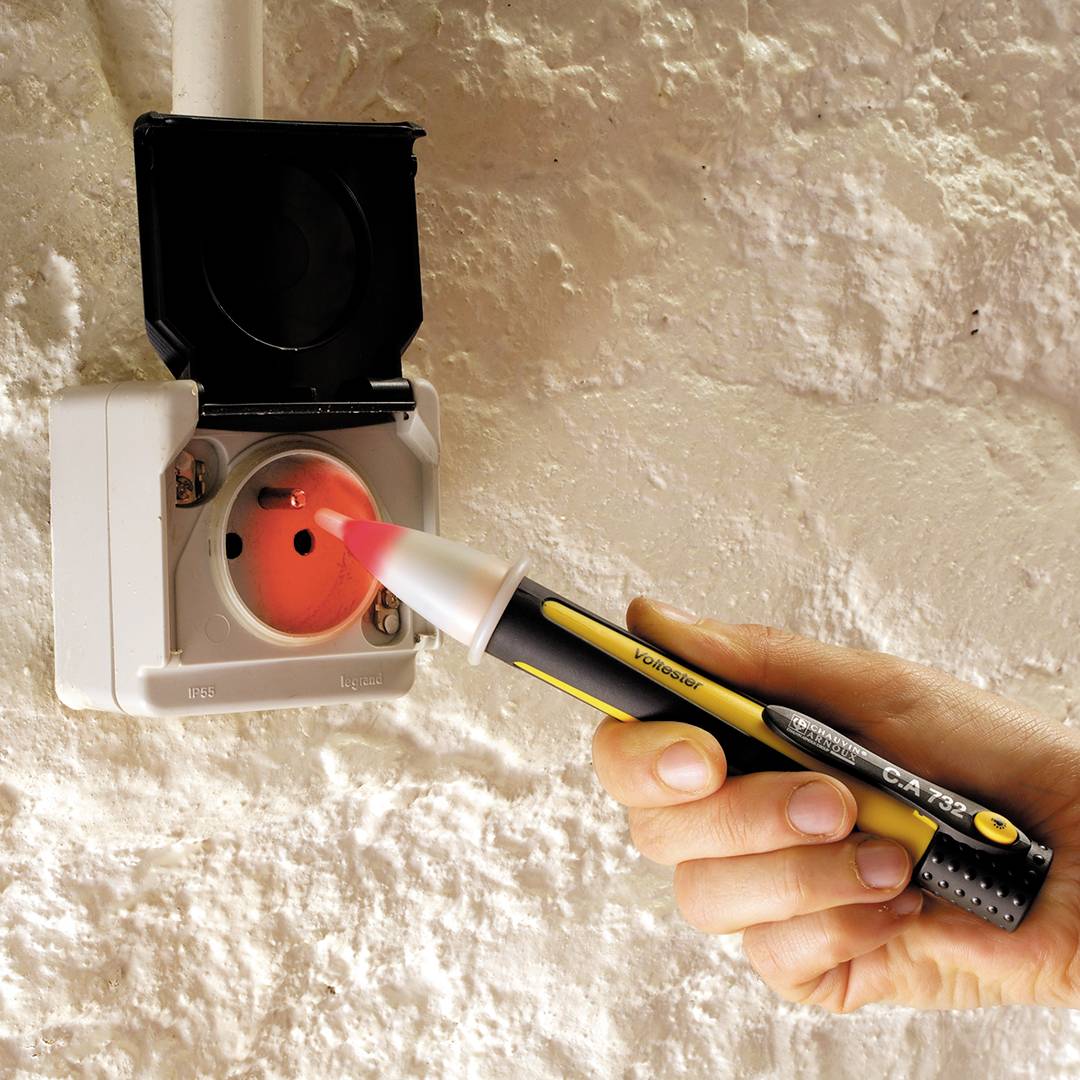 Une main tient un testeur de tension près d'une prise électrique ouverte sur un mur pour vérifier la tension.